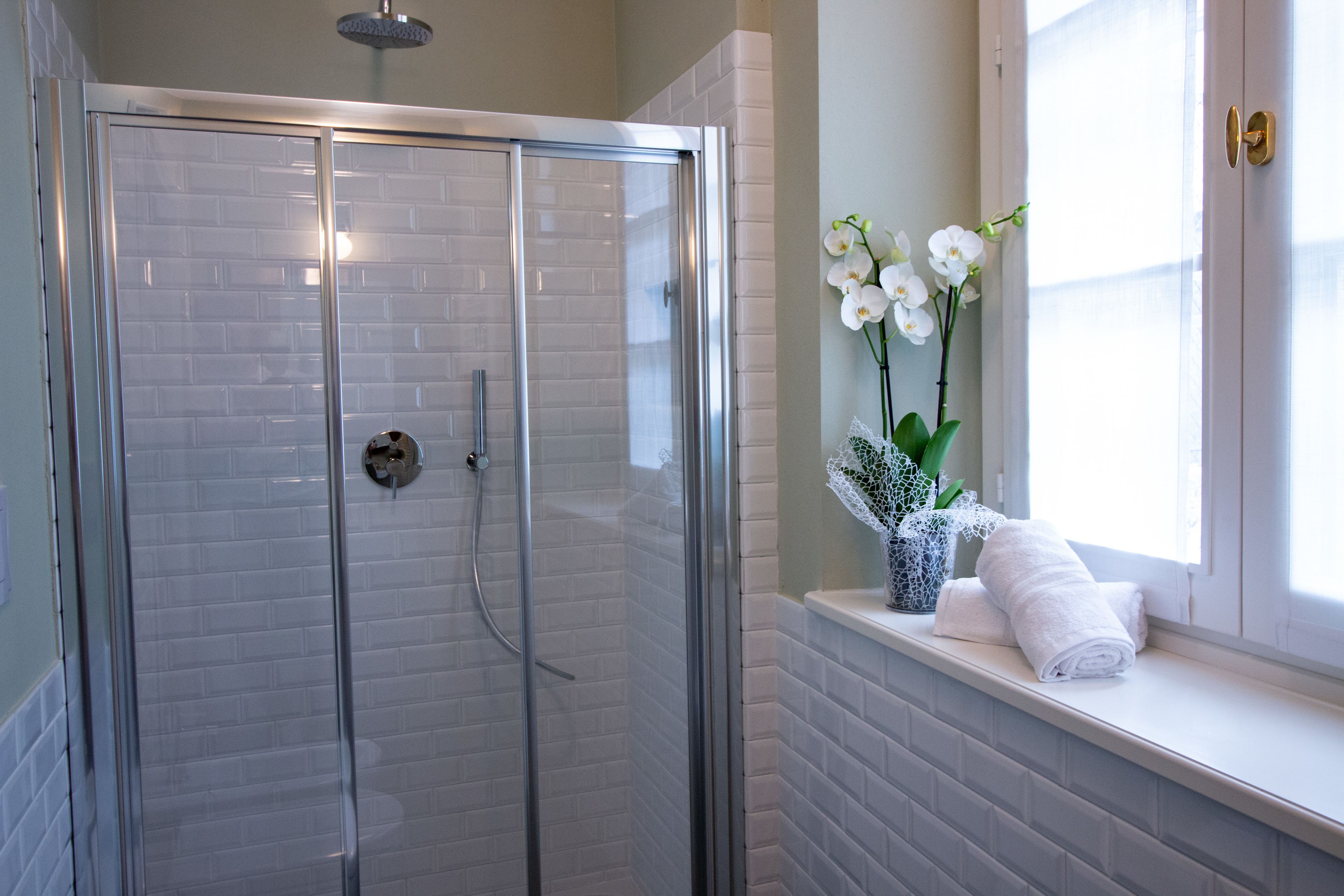 A glass-enclosed shower with white tiled walls, a window with white flowers, and a towel on the windowsill.