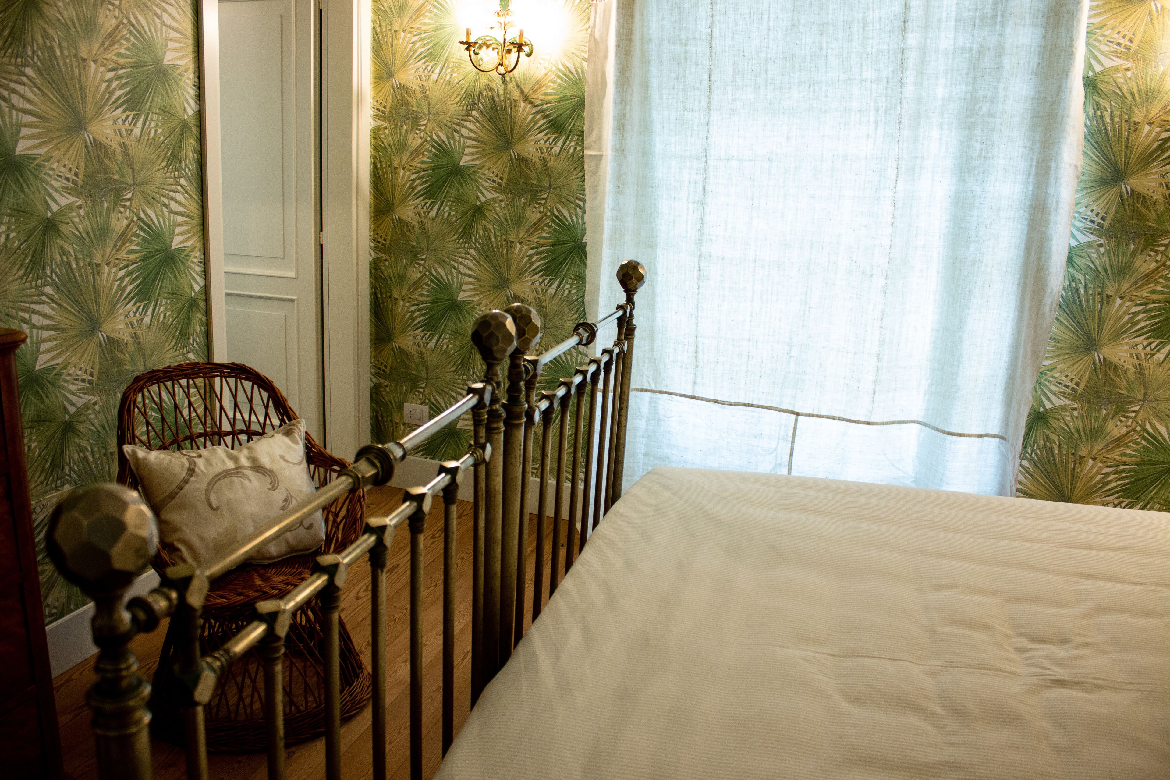 A room with a bed, a wicker chair with a cushion, a wooden railing, and a wall with a leafy pattern.