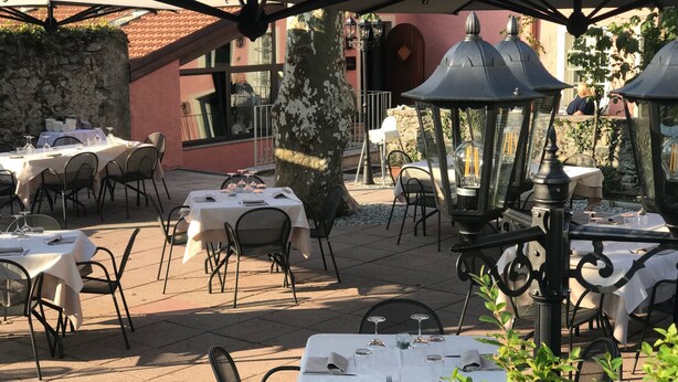 Un área de comedor al aire libre con mesas preparadas para dos, cubierta por sombrillas y rodeada de muros de piedra y follaje.