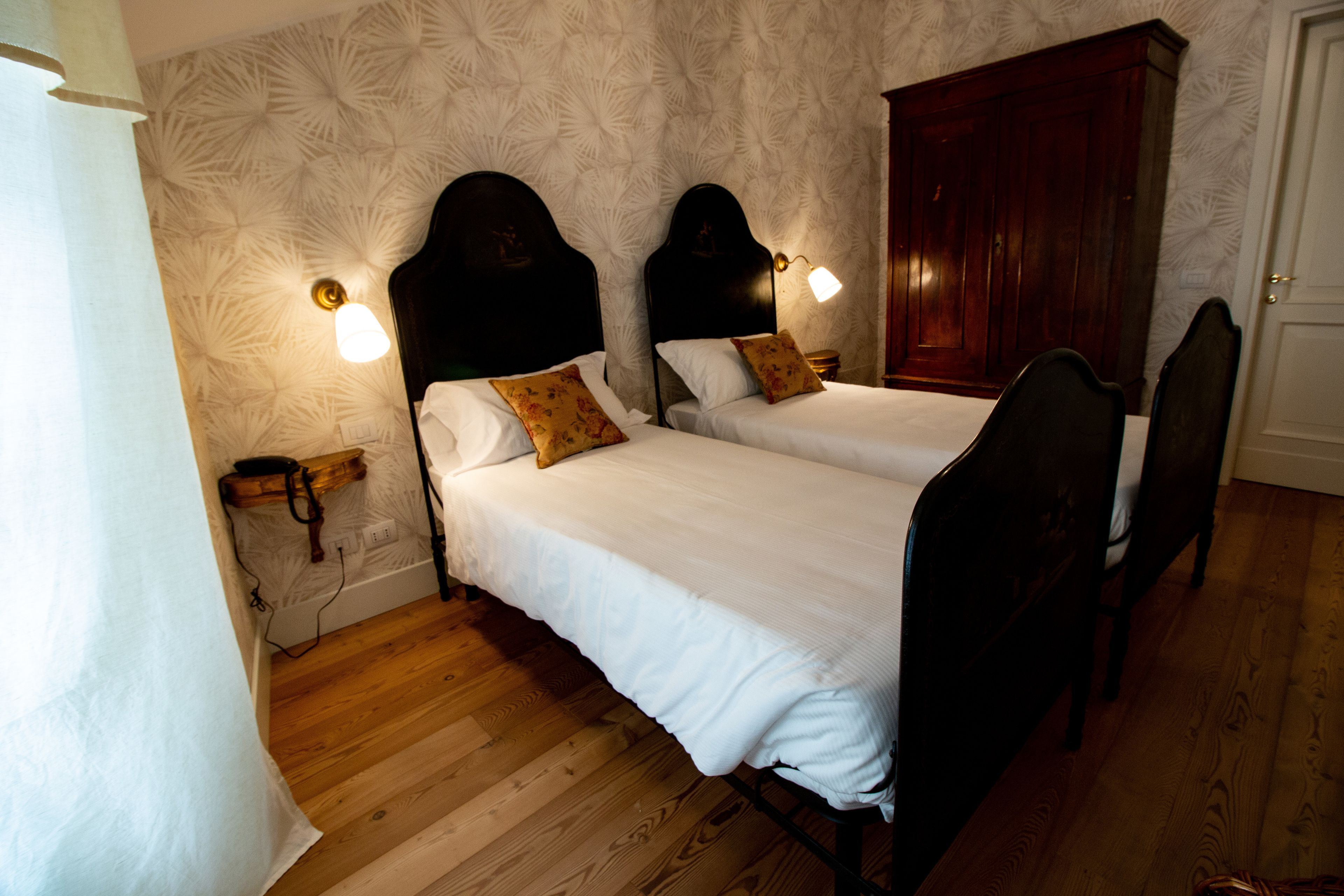 A hotel room with two beds, wooden flooring, a wardrobe, and wall-mounted lamps.
