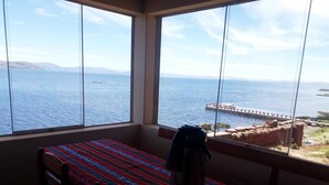 Une pièce dotée d’une grande fenêtre donnant sur la mer et un quai. On y trouve une table recouverte d’une nappe colorée et un récipient en métal posé dessus.