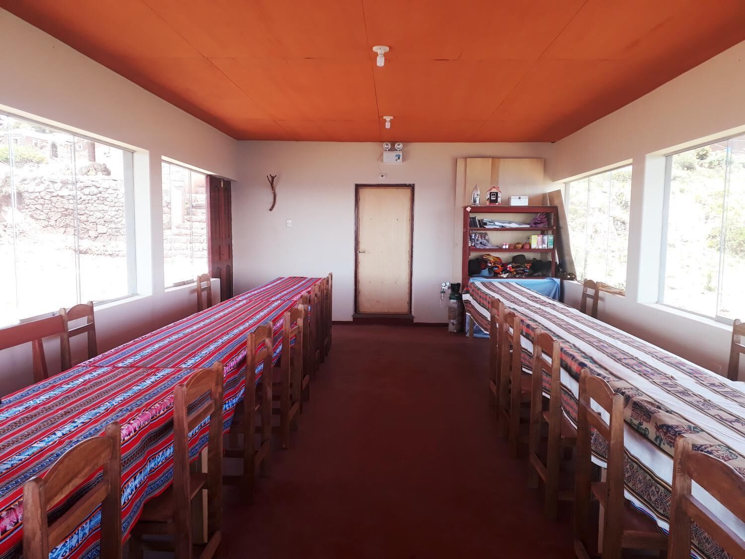 A dining area with a long table and chairs, a door, and a shelf with various items.