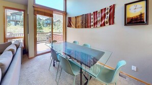A room with a glass table and chairs, a sofa, a large window, and a wall with a patterned tapestry and a framed picture. - Snowcreek Resort Vacation Rental 950 (Mammoth Lakes)