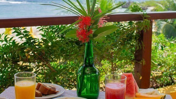 A table set with breakfast items including toast, fruit, and a green bottle, overlooking a scenic ocean view. - Pousada Funny (Cairu)