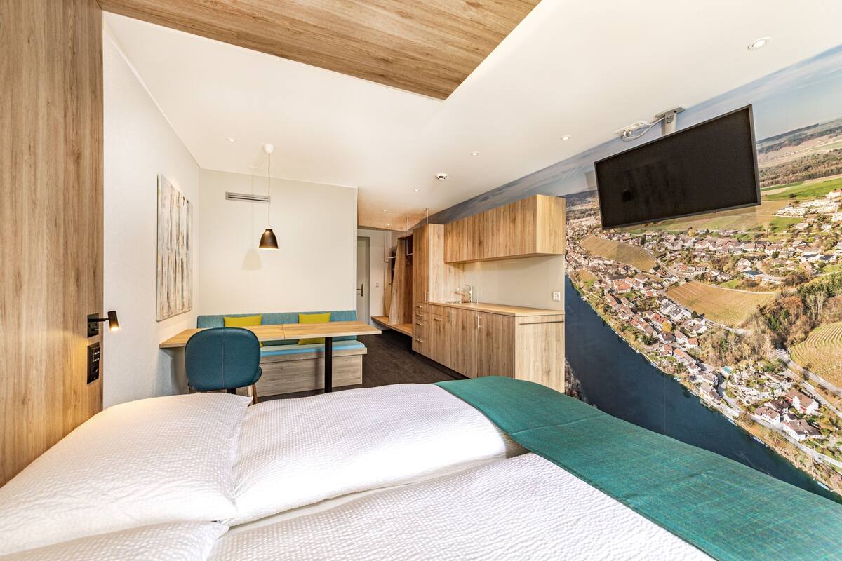 A modern hotel room with a large bed, a desk with a chair, a TV mounted on the wall, and a scenic mural of a landscape.