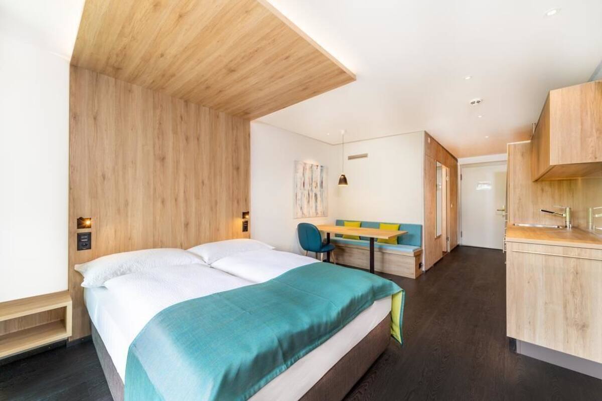 A modern hotel room with a large bed, a desk, and a kitchenette.
