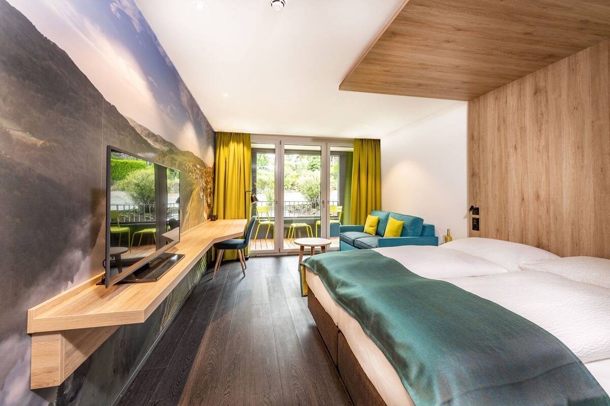 A modern hotel room with a large bed, a desk with a flat-screen TV, a sofa, and a balcony with yellow curtains.