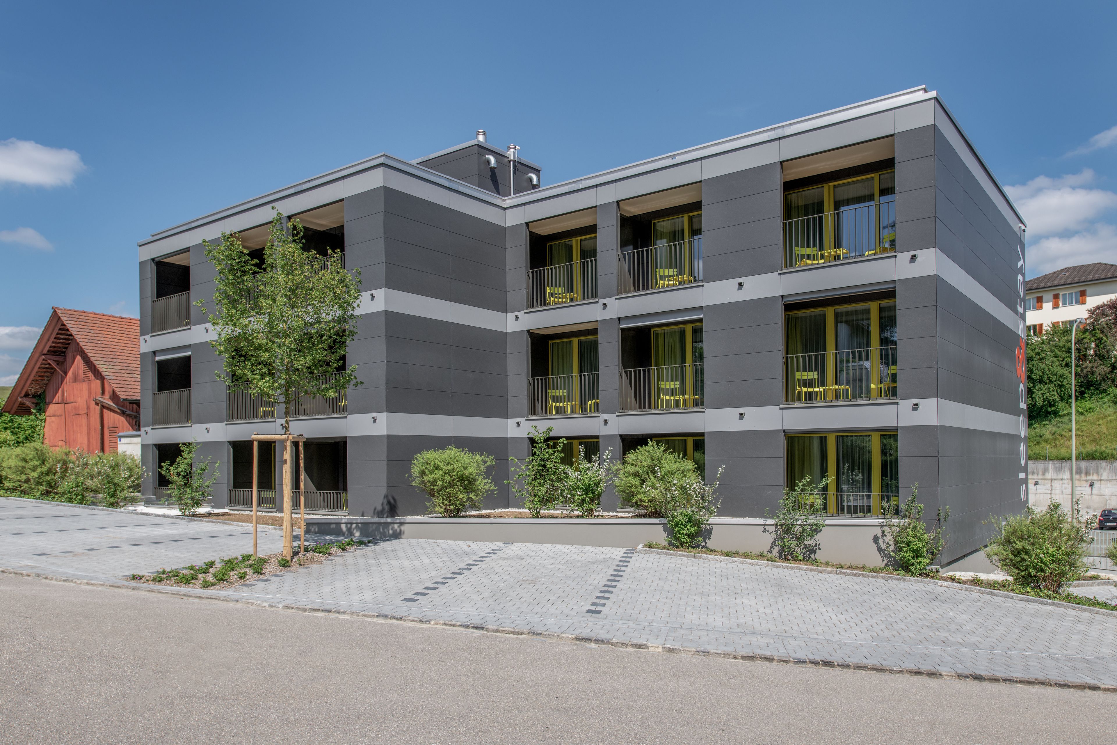 Un edificio moderno a più piani con balconi, un vialetto lastricato e alberi da giardino.