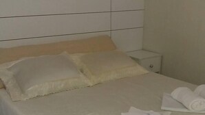 A neatly made bed with white linens and pillows, a small bedside table with a lamp, and a wall-mounted shelf. - Pousada Porto Brasil (Pocoes)