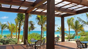 A covered outdoor area with wooden pillars, tables, and chairs, overlooking a beach with palm trees and the ocean. - Catalonia Grand Costa Mujeres All Suites & Spa - All Inclusive (Costa Mujeres)