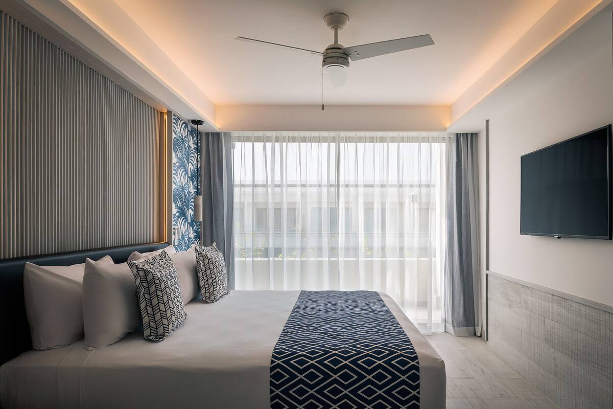 Ein modernes Hotelzimmer mit einem großen Bett, einem Deckenventilator, einem an der Wand montierten Fernseher und einem großen Fenster mit durchsichtigen Vorhängen.