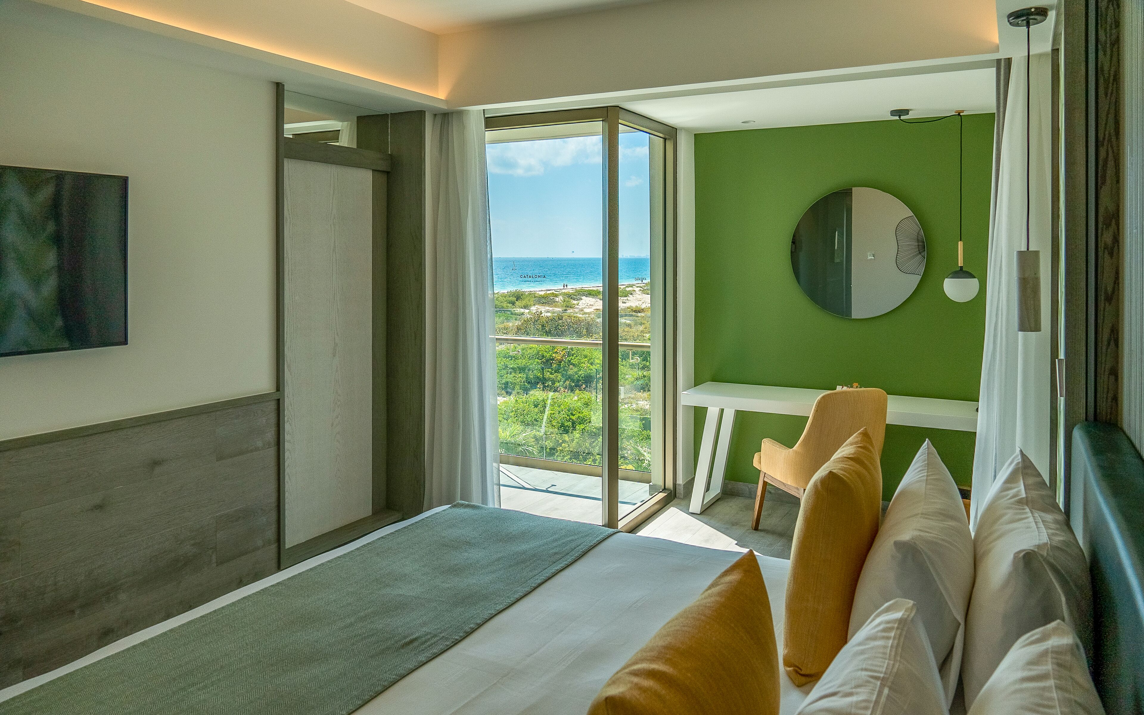 A bedroom with a large bed, a view of the sea, a green wall, and a round mirror.