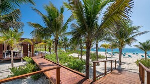 A wooden deck leading to a beach area with palm trees, lounge chairs, and a pool area. - Catalonia Grand Costa Mujeres All Suites & Spa - All Inclusive (Costa Mujeres)