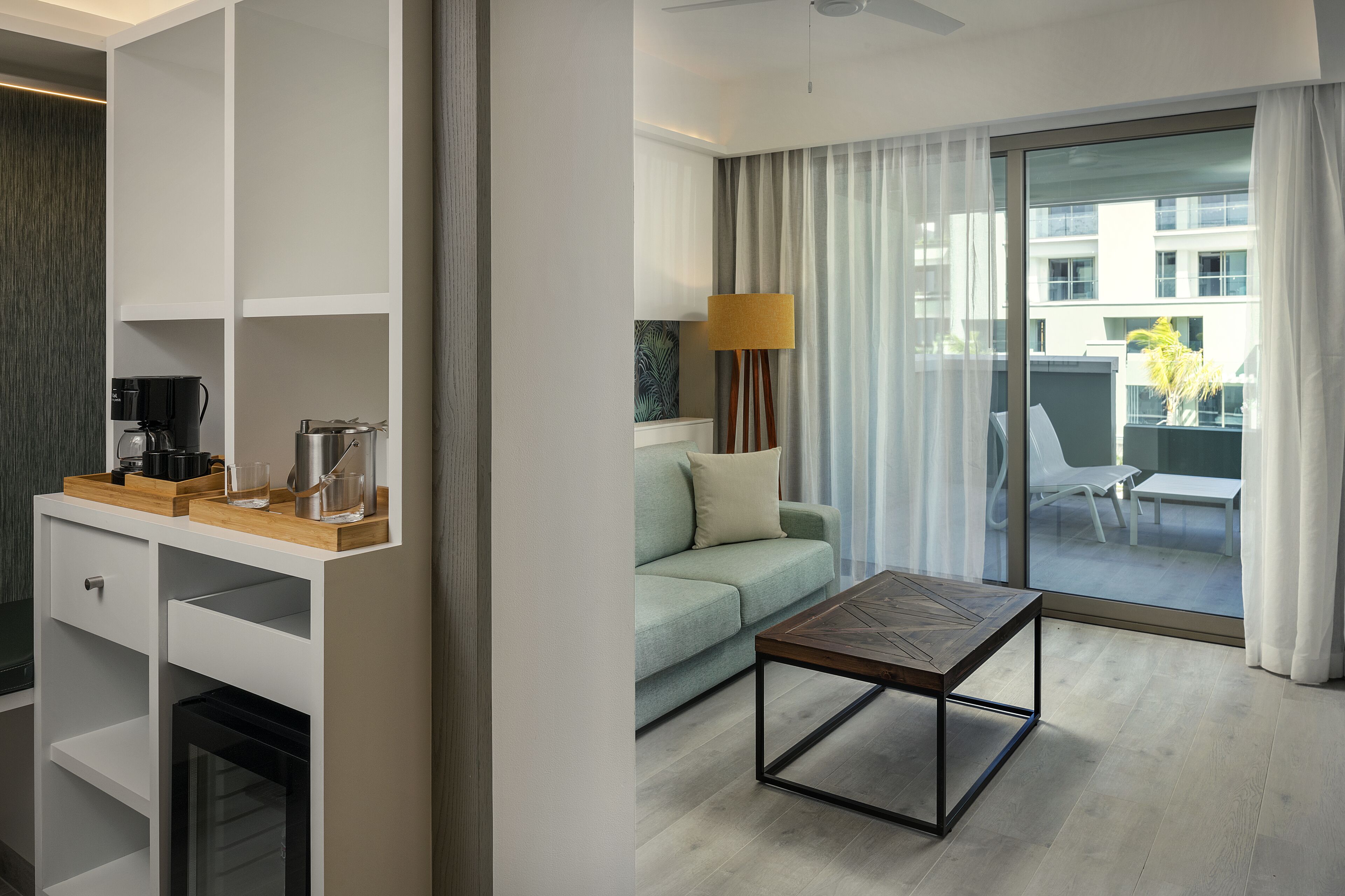 A modern hotel room with a sofa, a coffee table, a lamp, and a view of the outside balcony.