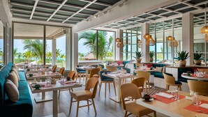 A restaurant with tables and chairs, large windows, and a view of palm trees. - Catalonia Grand Costa Mujeres All Suites & Spa - All Inclusive (Costa Mujeres)