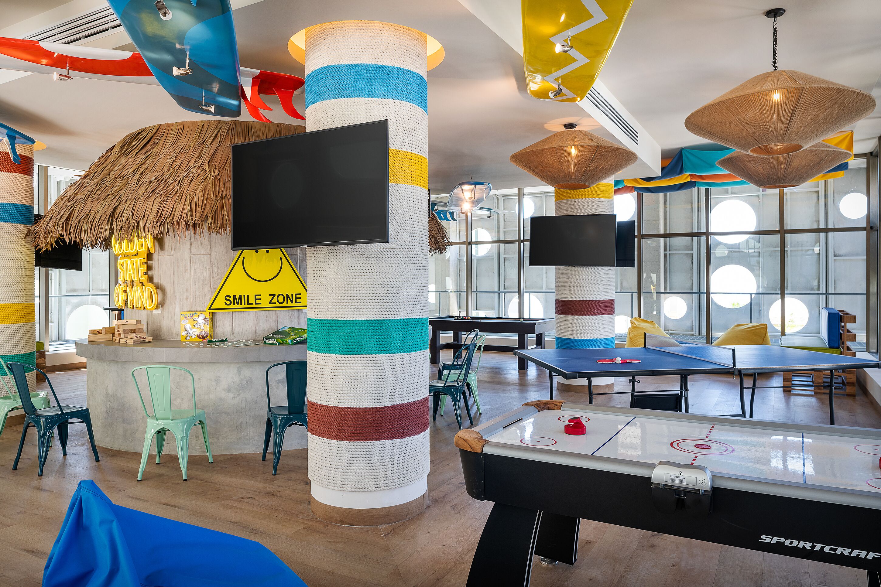 A brightly lit indoor space with a foosball table, striped columns, and a bar area with a neon sign.