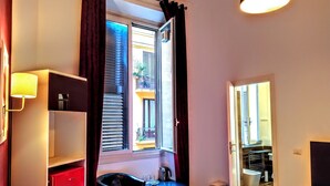 A hotel room with a bed, a desk, a chair, a red wardrobe, a window with curtains, and a bathroom visible through an open door. - NIK King Rooms (Rome)