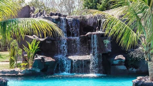 Un élément décoratif aquatique d’inspiration naturelle, avec de l’eau en cascade sur des rochers, le tout entouré d’une végétation tropicale.