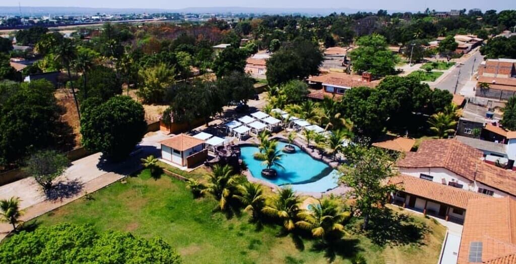Vista aérea de um resort com uma piscina central, rodeado por palmeiras e edifícios com telhados de terracota.