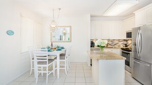Une cuisine lumineuse avec des meubles blancs, une table à manger avec des chaises, un lustre et un réfrigérateur.