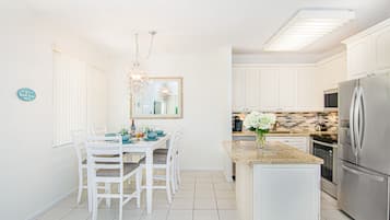 A bright kitchen with white cabinets, a dining table with chairs, a chandelier, and a refrigerator.