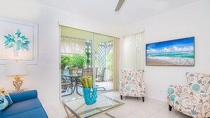 Un salon avec une table basse en verre, un canapé bleu, des fauteuils à motifs, un tableau encadré et, à travers une porte vitrée, une vue sur un espace extérieur aménagé avec une table et des chaises.