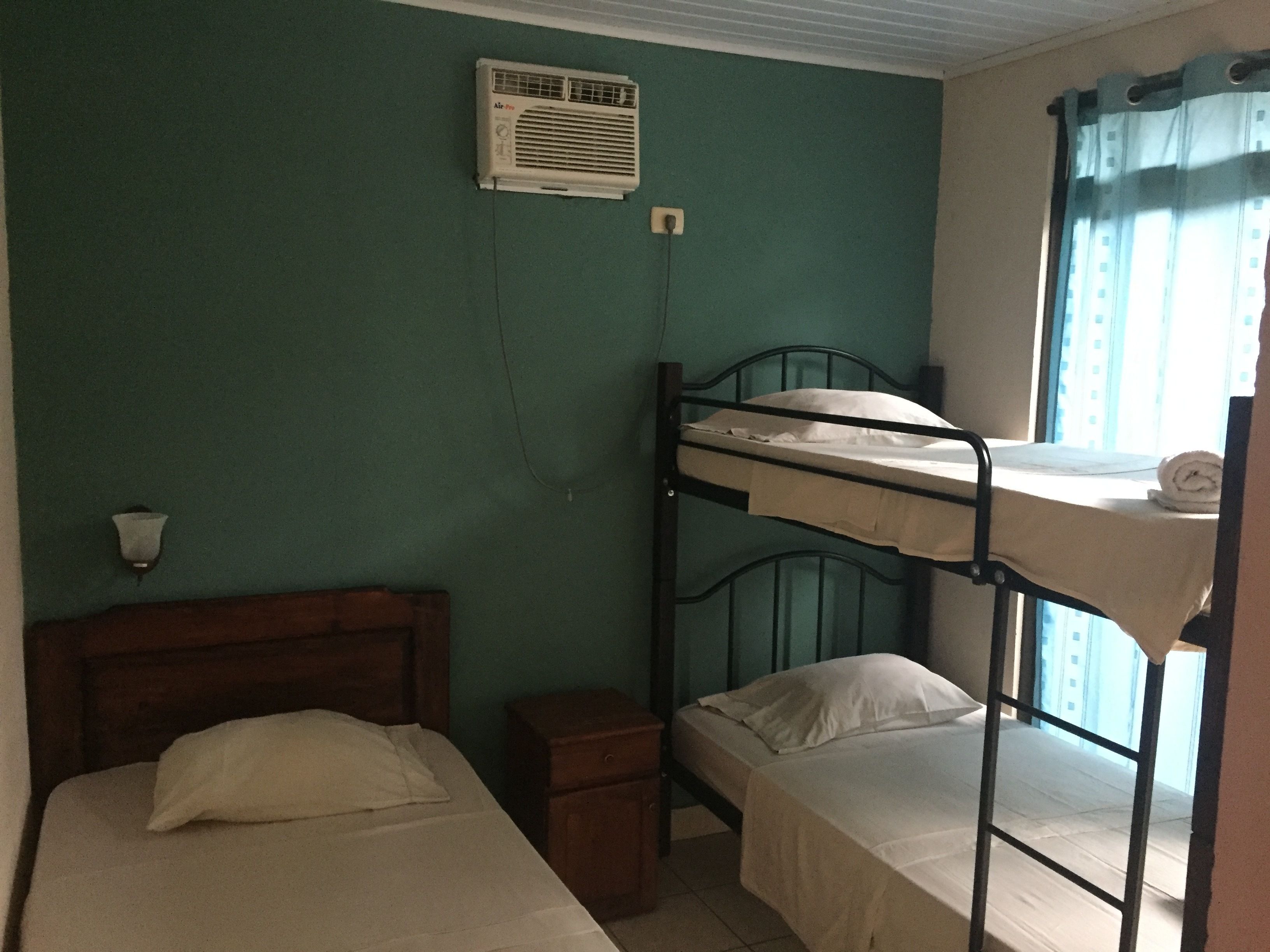 Quarto em dormitório com beliches, uma mesinha de cabeceira, ar condicionado fixado na parede e uma janela com cortinas.