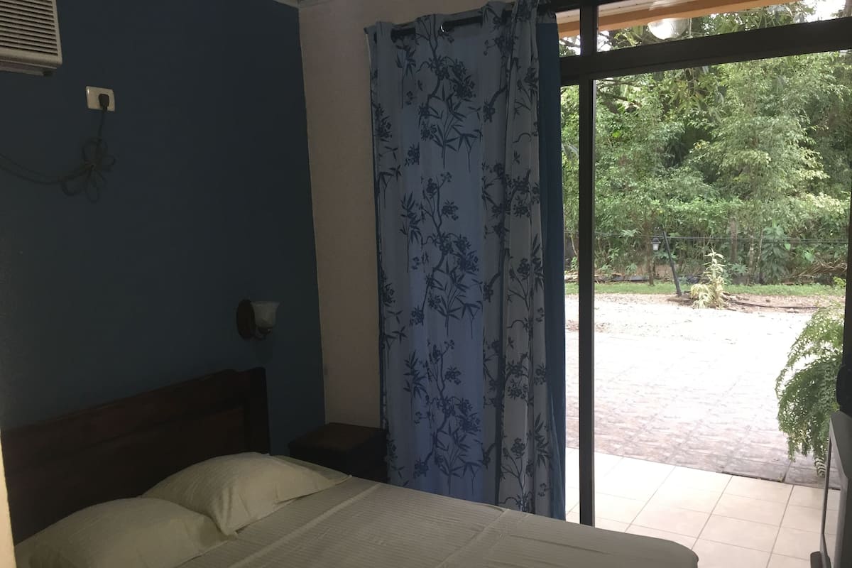 A bedroom with a bed, a nightstand, a wall-mounted lamp, and a view of a garden through a window with a floral curtain.