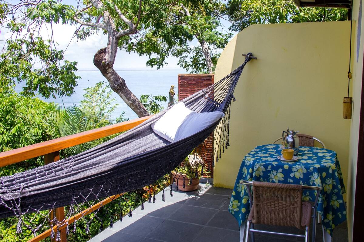Un hamac sur un balcon avec vue sur la mer, une table recouverte d’une nappe à motifs floraux et une chaise.