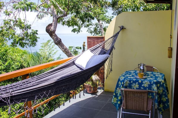 A hammock on a balcony with a view of the sea, a table with a floral tablecloth, and a chair.