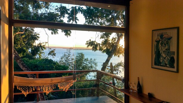 A room with a large window overlooking a body of water, a hammock, and a wooden shelf with a bottle and a glass. - Pousada VillaBahia (Cairu)