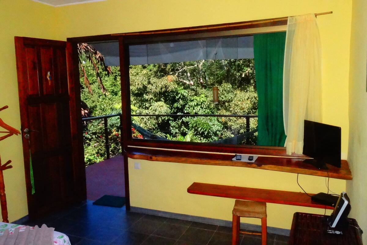 Une chambre avec une fenêtre donnant sur un jardin, un bureau avec une chaise et un lit muni d’un rideau.