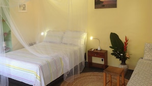 A bedroom with a bed covered by a mosquito net, a bedside table with a lamp, a potted plant, and a painting on the wall. - Pousada VillaBahia (Cairu)