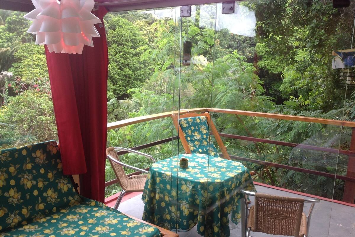 Un balcon agrémenté de meubles en osier, une table recouverte d’une nappe à motifs floraux et une vue sur une végétation luxuriante.