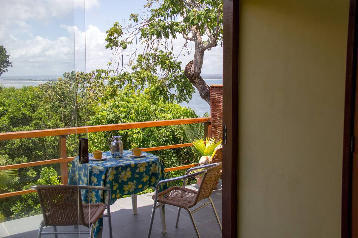 Un balcon avec une table dressée pour deux personnes, offrant une vue sur un paysage luxuriant et une étendue d’eau.