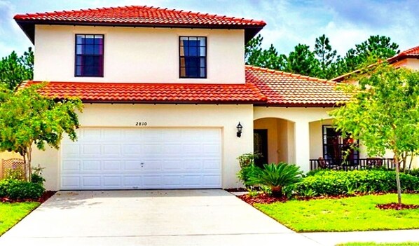 A two-story house with a red-tiled roof, a white garage door, and a well-maintained front yard with shrubs and a small tree. - 6 miles to WDW: 4 Bedroom/3+ Bath, Private Pool/Hot Tub, Wifi, Game Room, Clean (Kissimmee)