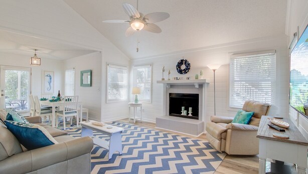 A spacious living room with a fireplace, a ceiling fan, and a dining area with a table and chairs. - 72 Pelican Bay | AvantStay | Community Pool, Decks (Isle of Palms)