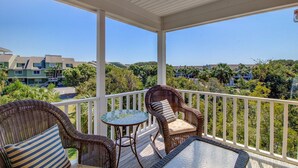 A covered balcony with wicker furniture and a mosaic table, overlooking a residential area with multiple buildings and greenery. - 72 Pelican Bay | AvantStay | Community Pool, Decks (Isle of Palms)