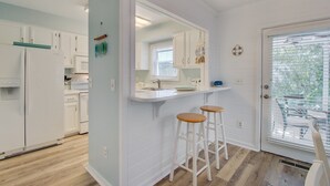A modern kitchen with white cabinets, a central island with stools, and a large window with blinds. - 72 Pelican Bay | AvantStay | Community Pool, Decks (Isle of Palms)