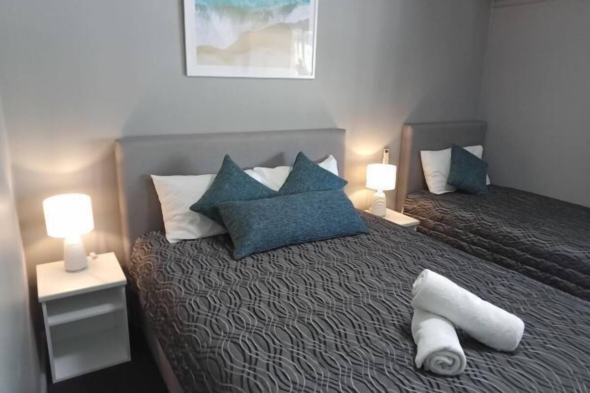 A hotel room with two beds, each with a headboard, bedside lamps, and a rolled towel on the bed.