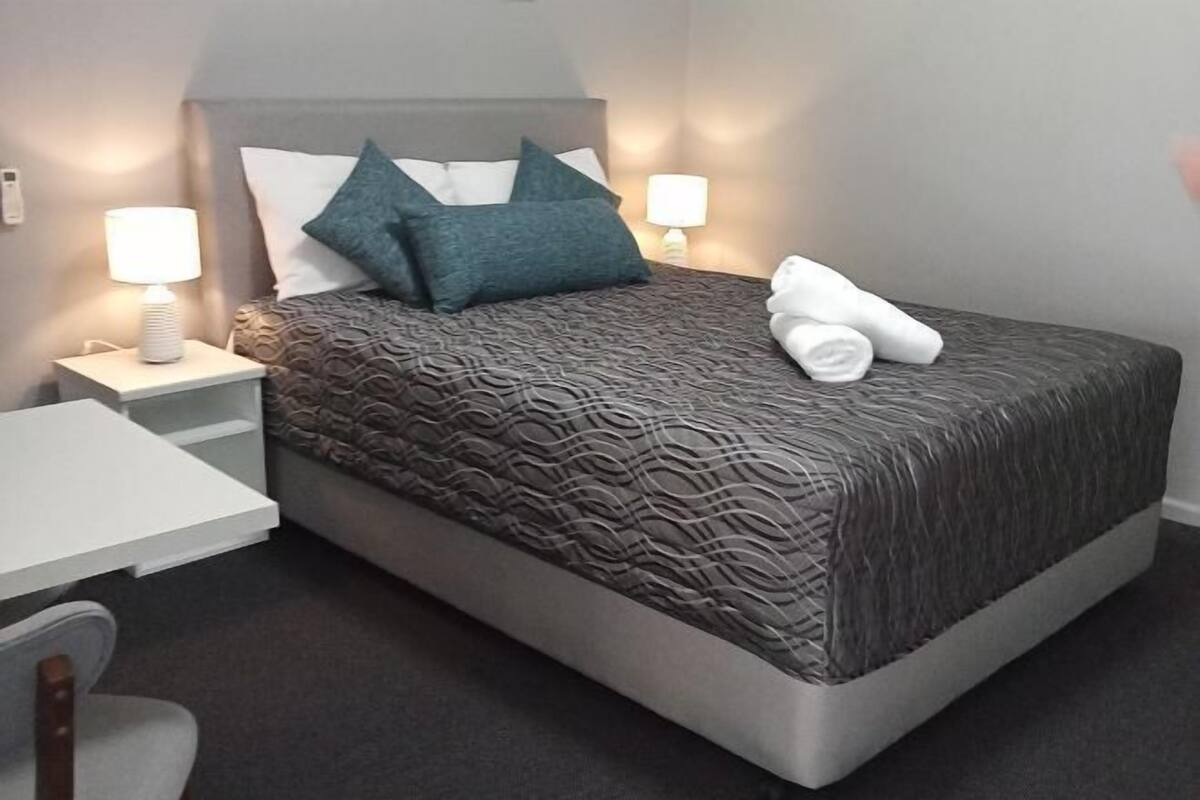 A neatly made bed with a patterned bedspread, two pillows, and a rolled towel on the side. There is a small bedside table with a lamp and a chair in the corner.