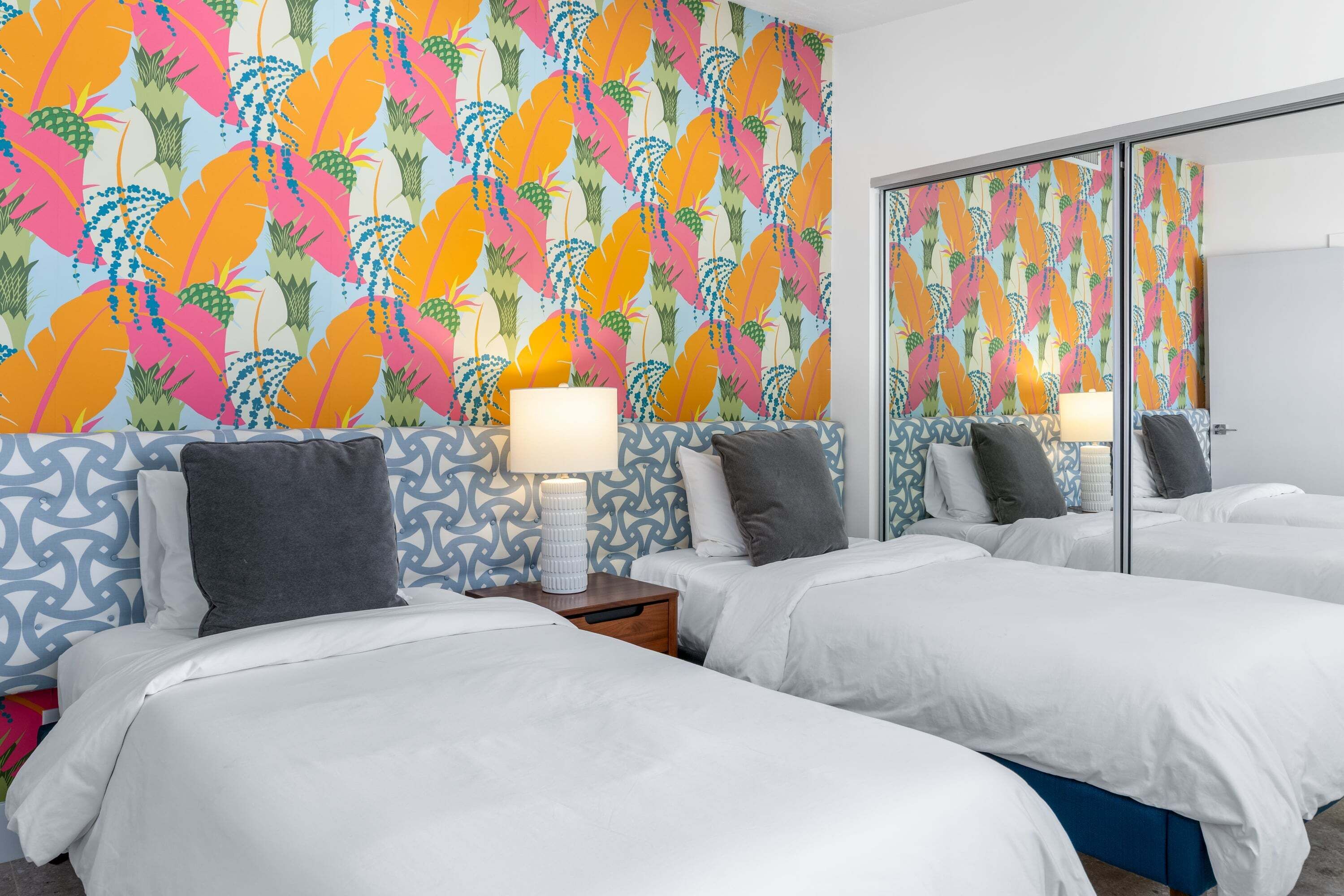 A hotel room with a mirrored closet, two beds with white bedding, a patterned wall, and a bedside table with a lamp.