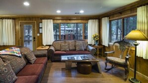 Una sala de estar acogedora con sofá, sillón y mesa de centro, paredes de madera y ventanas amplias con cortinas.