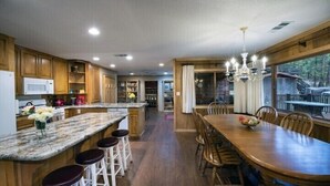 Una cocina con armarios de madera, encimera de granito, comedor con mesa y sillas de madera, y vista al exterior a través de grandes ventanas.