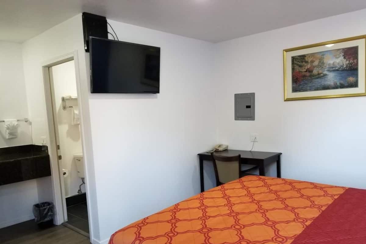 A hotel room with a bed, a desk, a chair, a television, a painting, and a bathroom visible through an open door.
