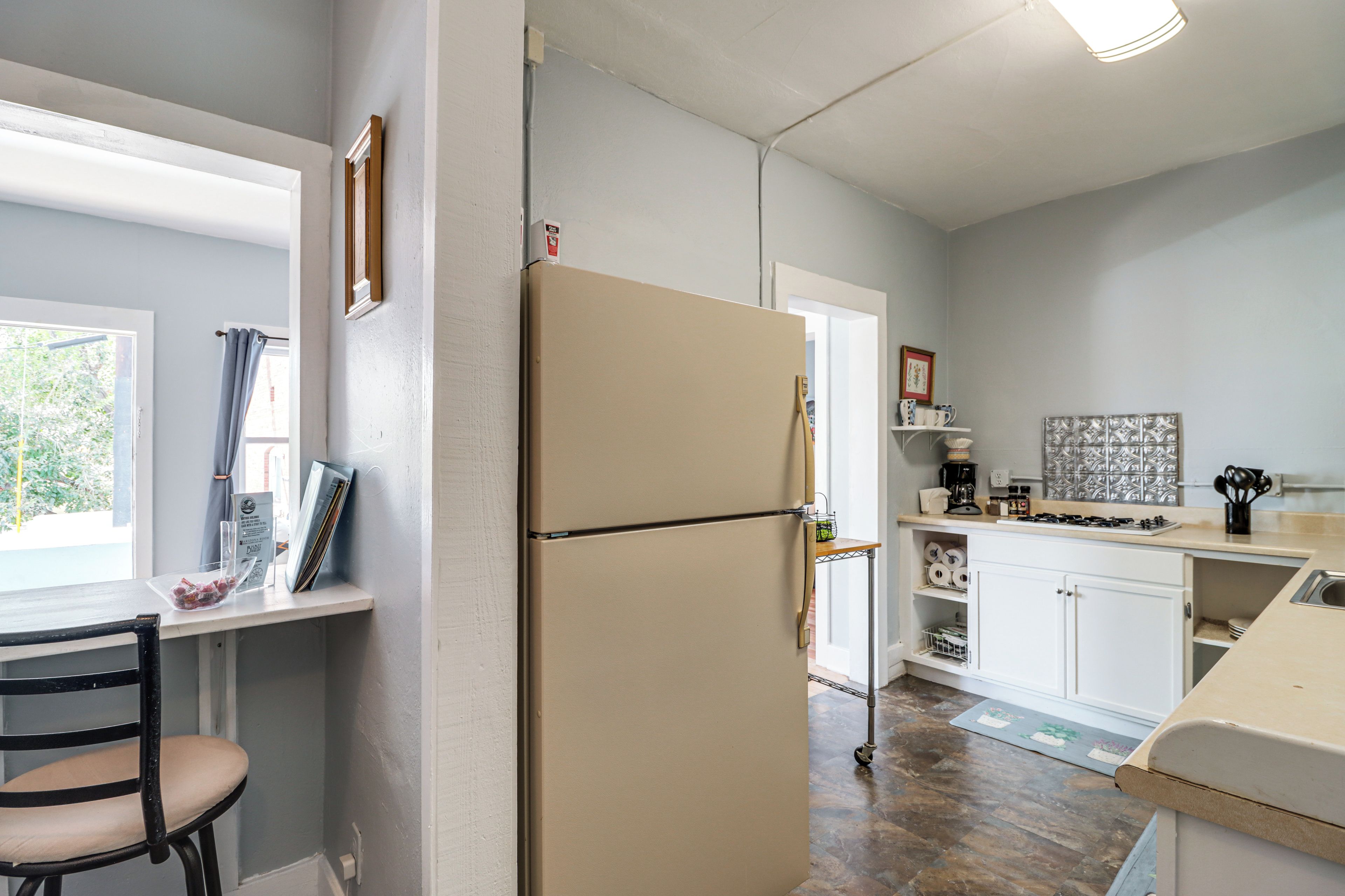 A compact kitchen with a refrigerator, a dining area with a chair, and a window with curtains.