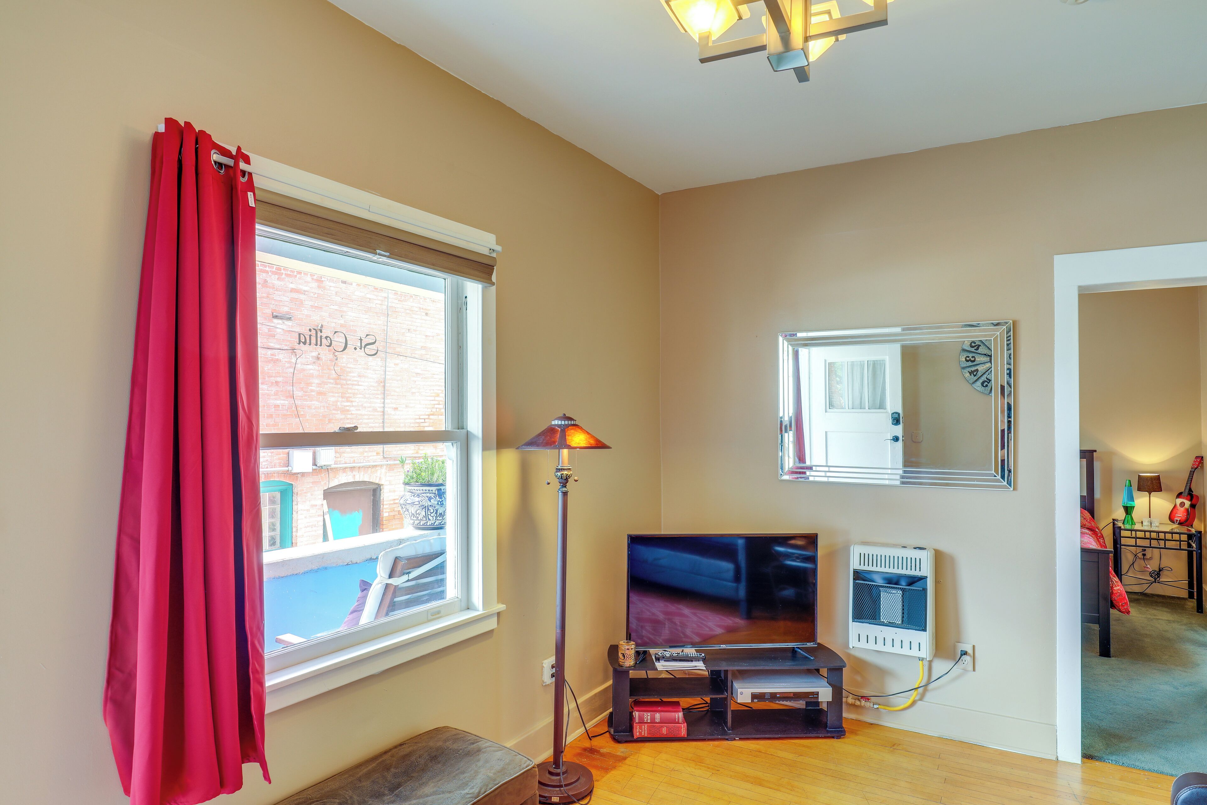 A living room with a TV, a floor lamp, a window with red curtains, a mirror, and a radiator.