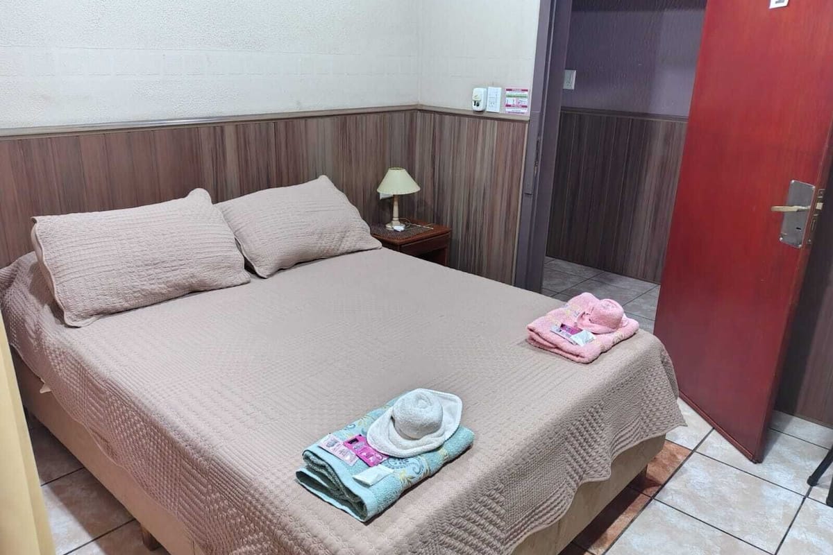 Uma cama de solteiro com um edredom bege, duas almofadas, uma mesa de cabeceira com abajur, uma porta com número e uma toalha na cama.