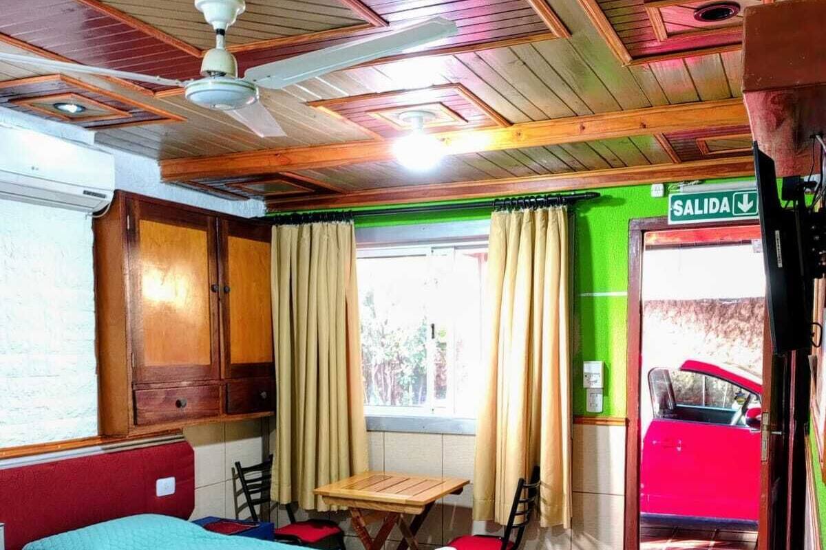 Quarto com duas camas, uma mesa, cadeiras, ventilador de teto, janela com cortinas e uma porta com a inscrição 'SALIDA'.