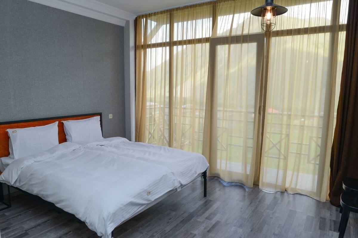 A bedroom with a large bed, a wooden headboard, and a view of a balcony through sheer curtains.